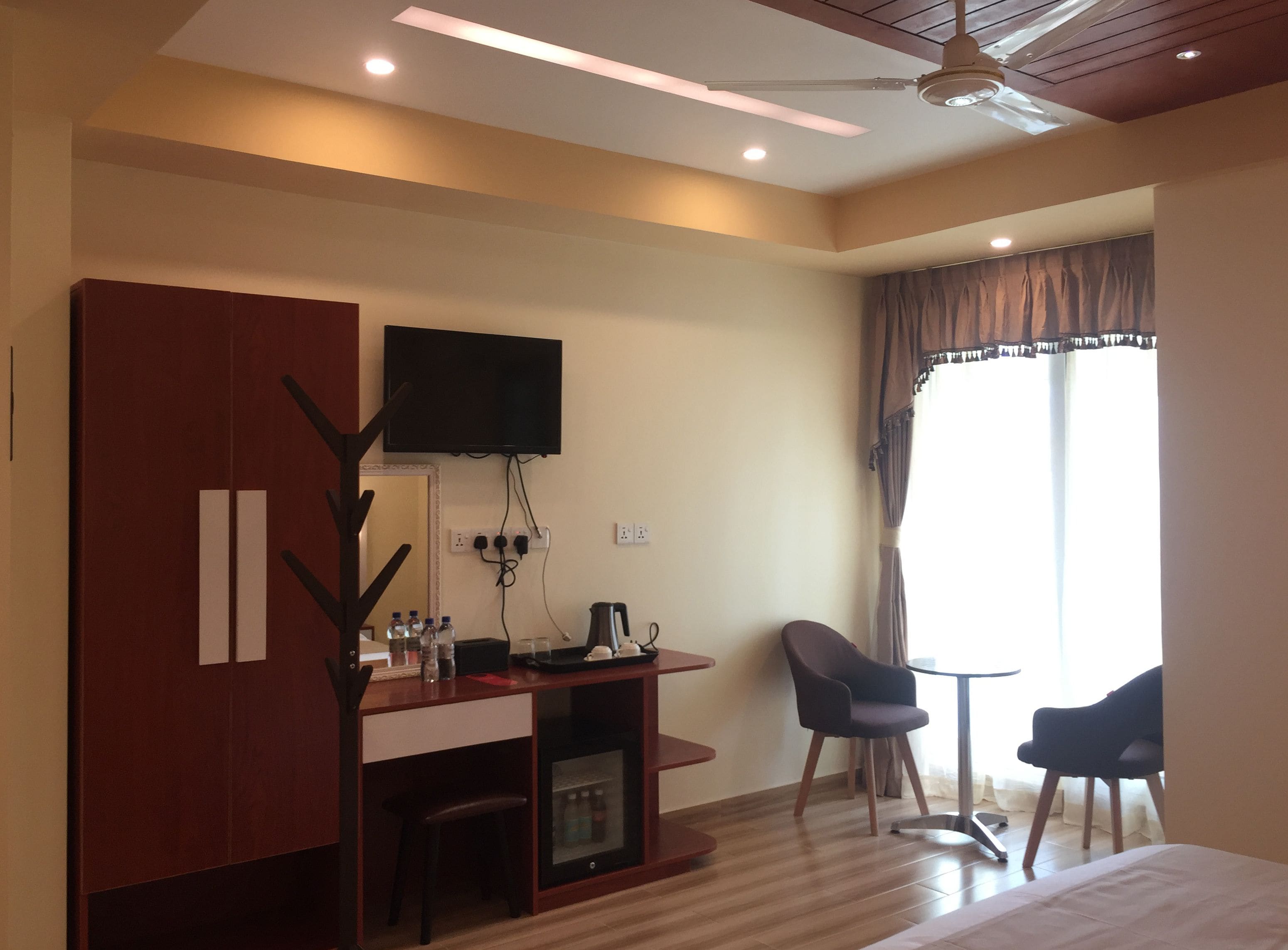 A hotel room with a wooden desk, a television mounted on the wall, a chair, a bed, and a window with curtains.