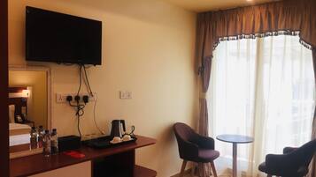 A hotel room with a flat-screen TV mounted on the wall, a desk with a kettle, cups, and a tray, two chairs, and a small table.