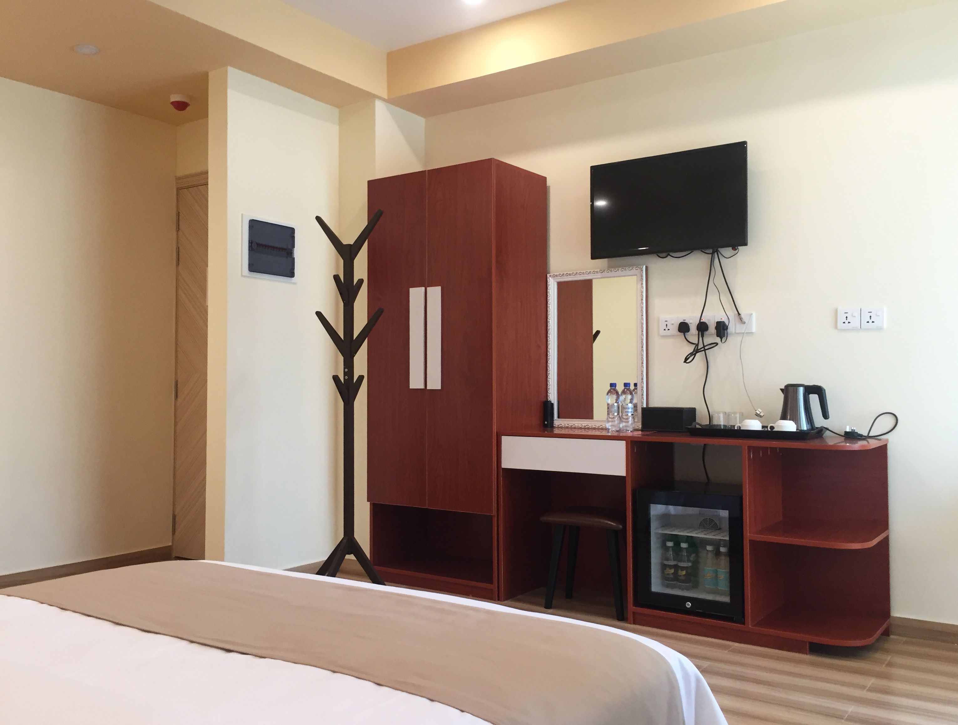 A hotel room with a bed, a desk with a mirror, a television, a small refrigerator, and a coat rack.