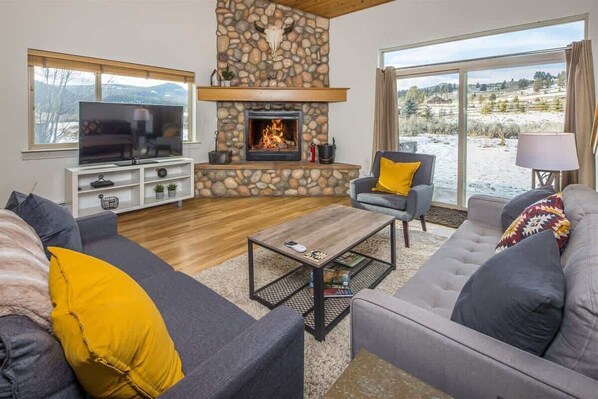 A living room with a stone fireplace, a television, sofas, and a view of a snowy landscape.