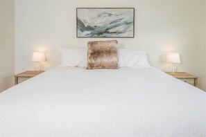 A neatly made bed with a white bedspread and two white pillows, a brown accent pillow, and a framed abstract painting above the bed.