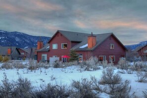 A red two-story house with a chimney, surrounded by snow and sparse vegetation.