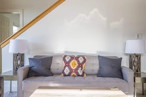 Una sala de estar moderna con un sofá gris, dos mesitas de noche con lámparas y un cojín decorativo estampado.