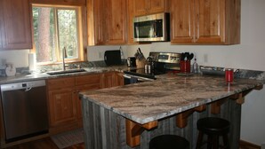 A kitchen with wooden cabinets, a granite countertop, a stainless steel refrigerator, and a window with a view of trees. - Luxury Guest House With River Access  (Bigfork)