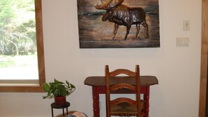 A room with a plaid sofa, a wooden dining table with a chair, a moose painting on the wall, and a window with a view of greenery. - Luxury Guest House With River Access  (Bigfork)
