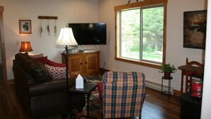 A living room with a couch, a television, a lamp, a window with a view of trees, and a fireplace. - Luxury Guest House With River Access  (Bigfork)