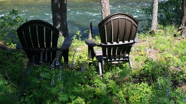 Two Adirondack chairs facing a river surrounded by dense forest. - Luxury Guest House With River Access  (Bigfork)