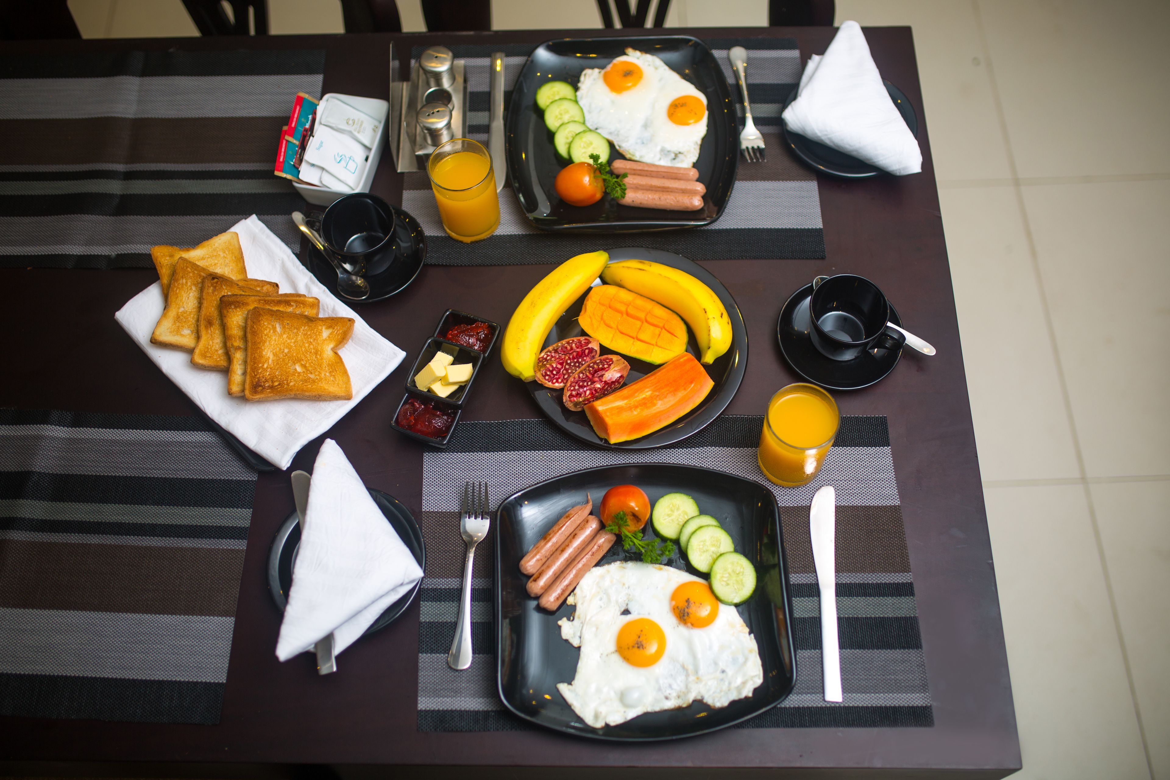 Uma mesa de café da manhã com pratos de ovos, linguiça, torradas e frutas, acompanhados de acompanhamentos e um copo de suco de laranja.