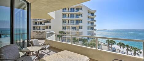 A balcony with a view of the ocean, furnished with chairs and a table, overlooking a beachfront building.