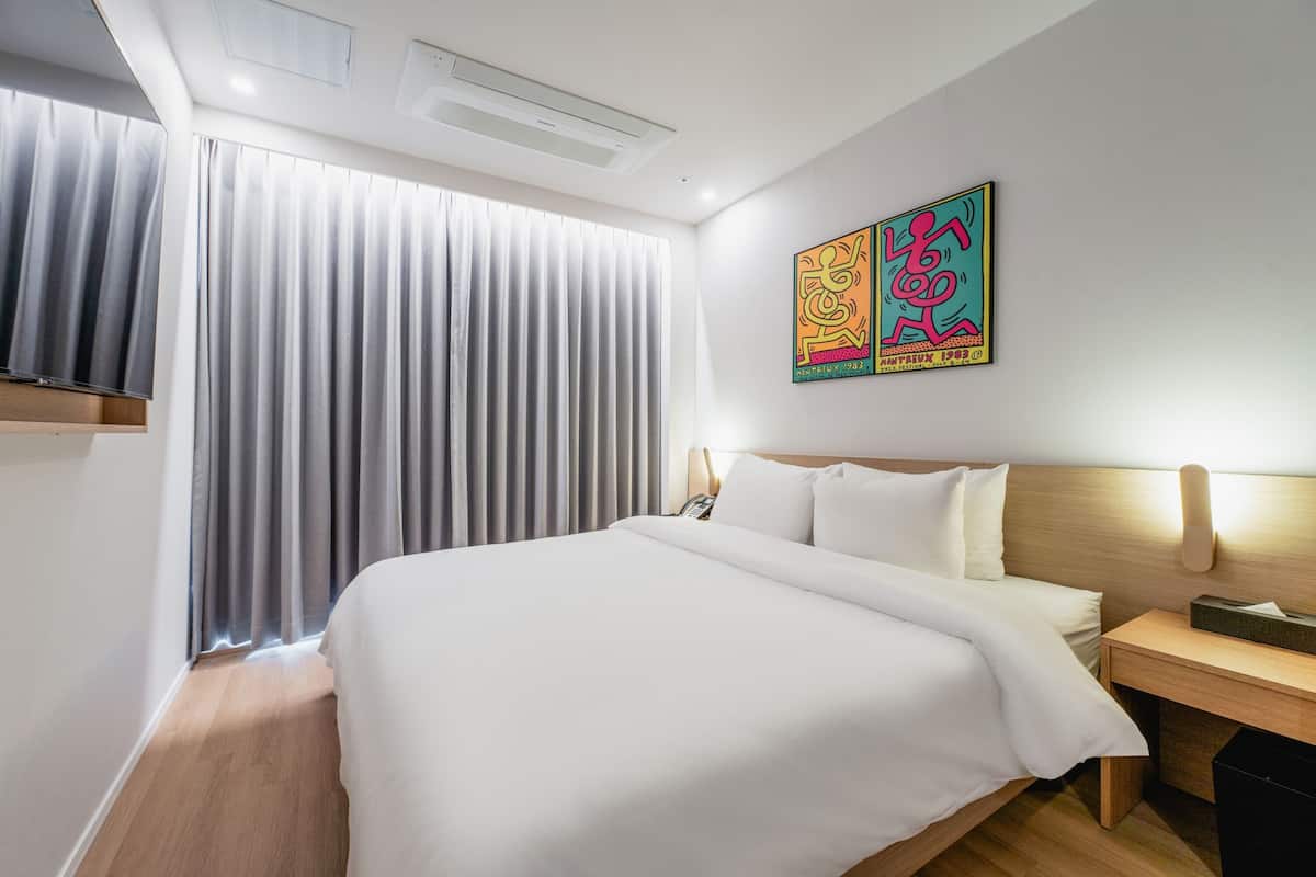 Hotellihuone, jossa on suuri sänky, puinen pääty, yöpöytä lampun kanssa ja ikkuna, jossa on verhot.