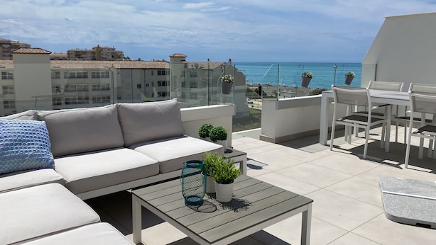 Une terrasse sur le toit, avec un canapé blanc, une petite table et un ensemble de mobilier de jardin. La terrasse offre une vue sur une étendue d’eau et sur des bâtiments en arrière-plan.