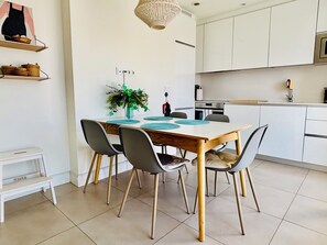 Et moderne køkken med hvide skabe, et spisebord med stole, en hængende lampe og en lille plante på væggen.