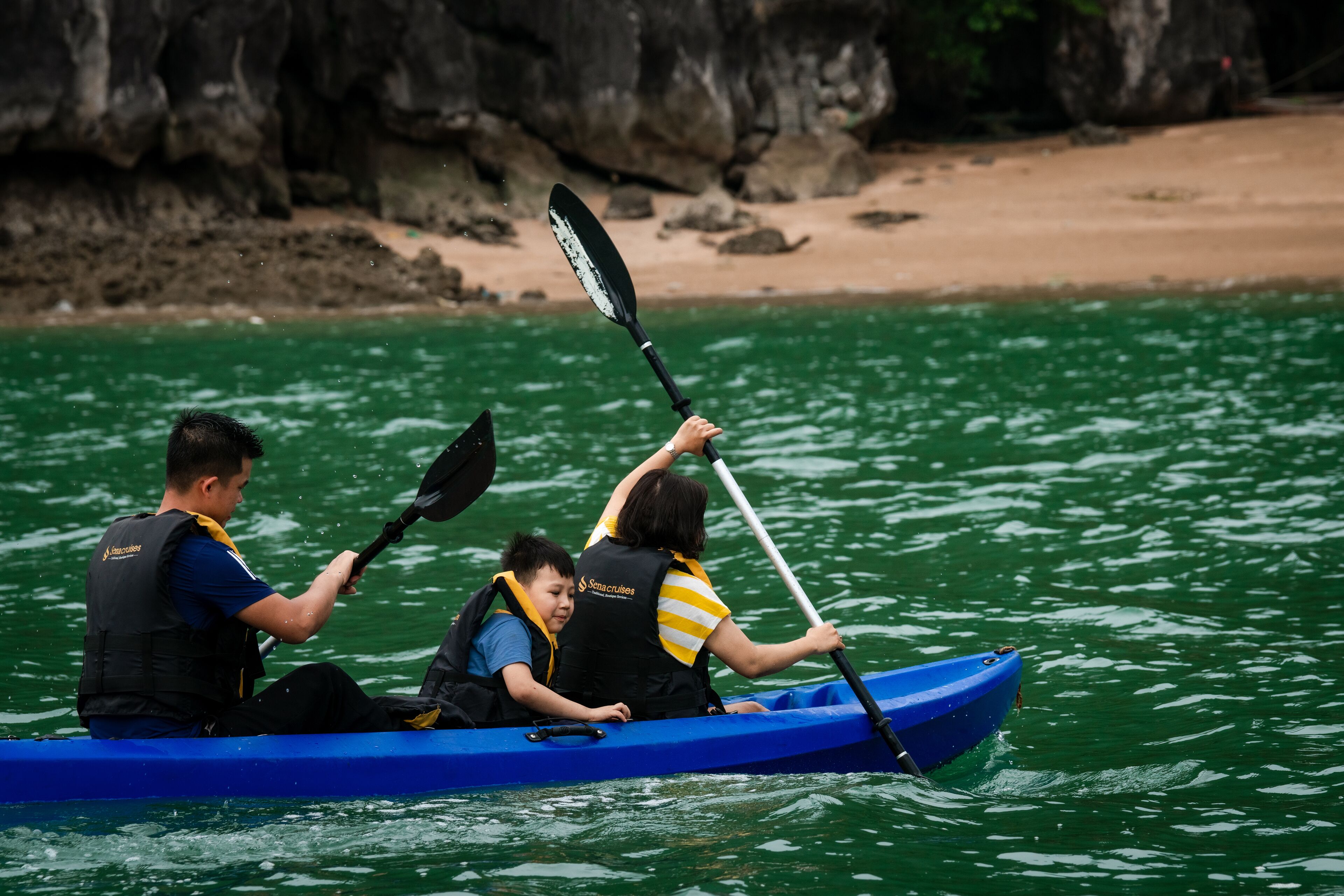 Three people in a blue kayak on a body of water with a rocky shoreline in the background.
