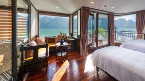 Quarto com uma grande janela que oferece vista para um lago e montanhas, uma cama, área de estar com mesa e uma varanda com grade.