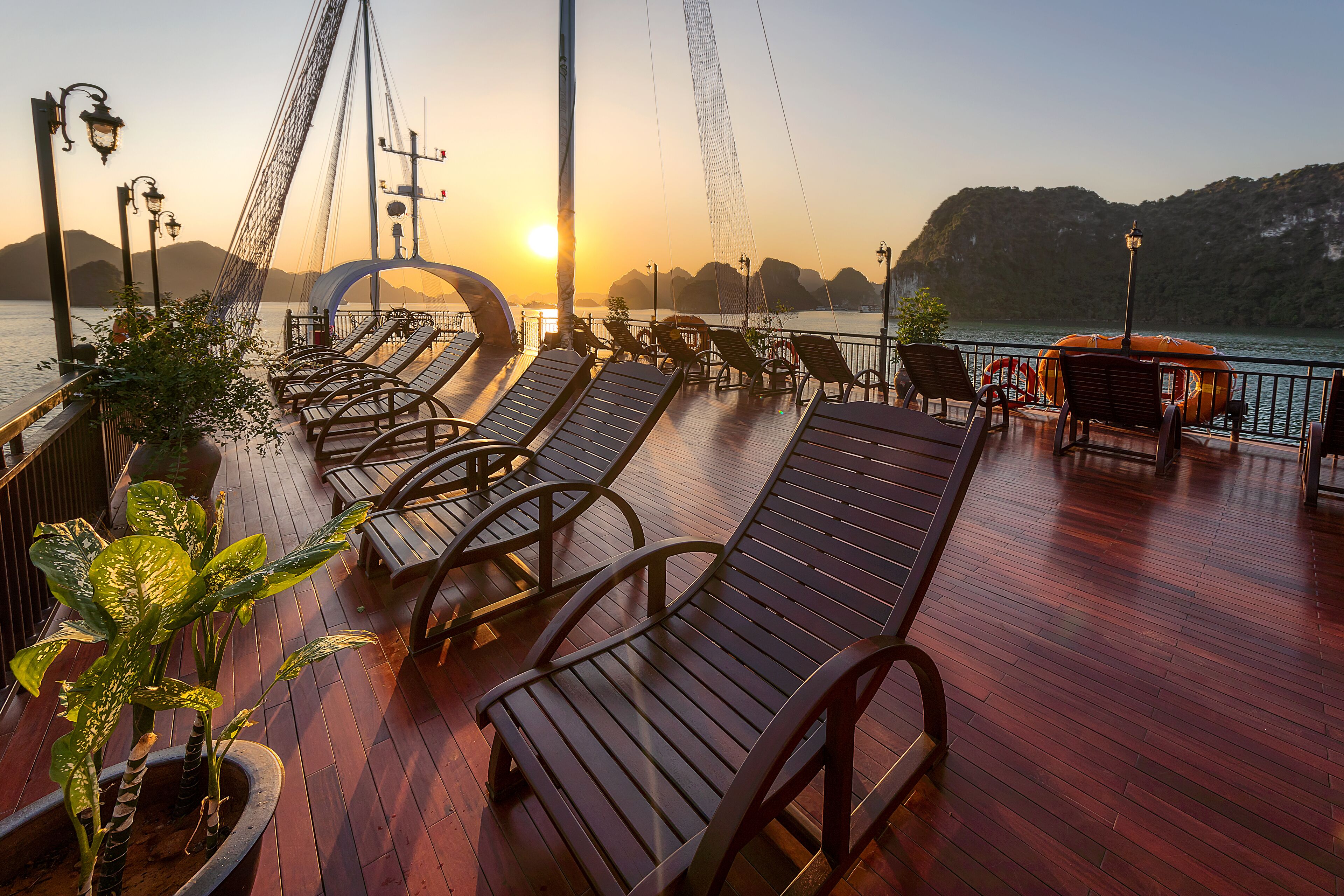 A deck with sun loungers, potted plants, and a view of the sunset over water.