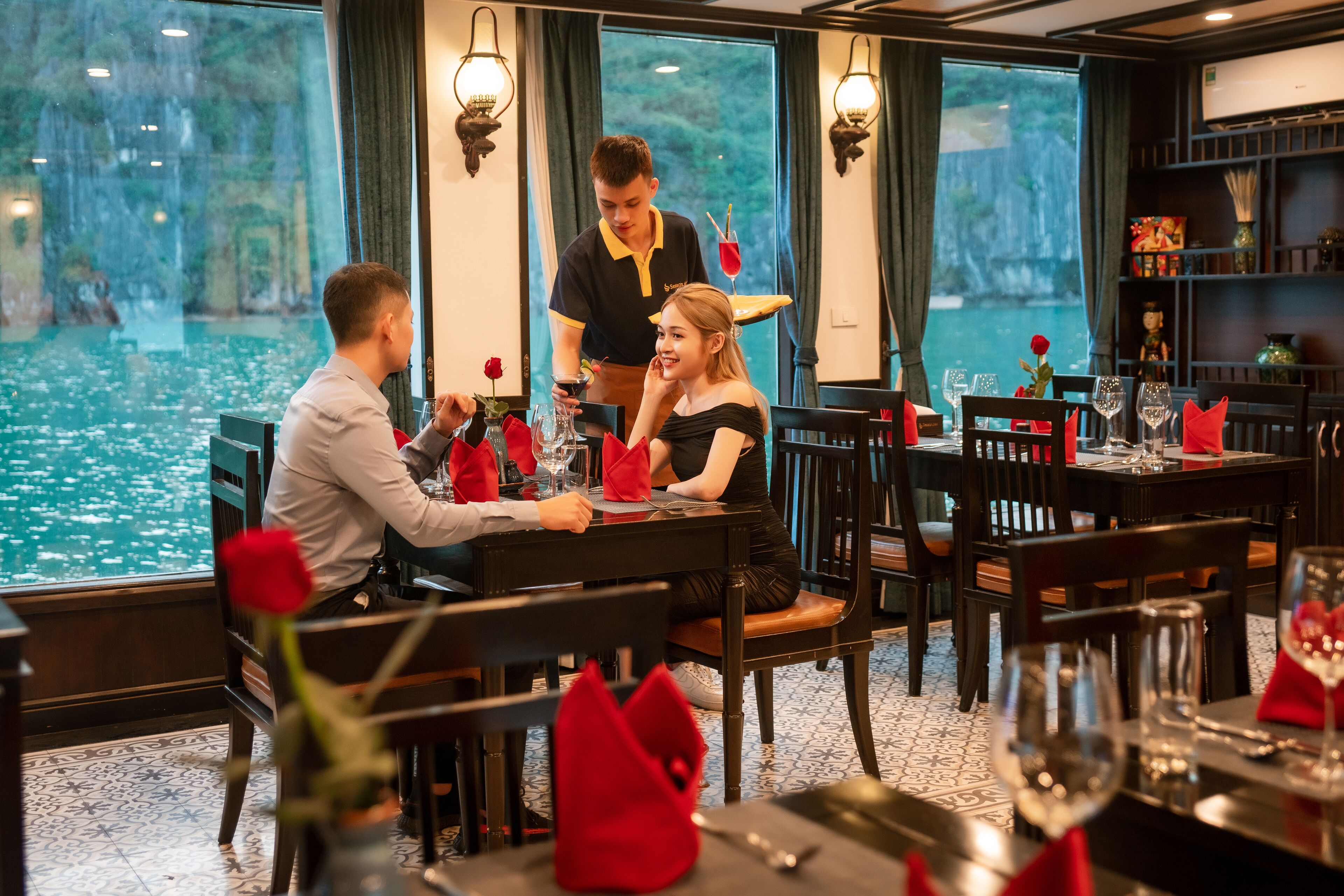 A restaurant with a view of a body of water, tables set with red napkins and glasses, and a waiter attending to guests.