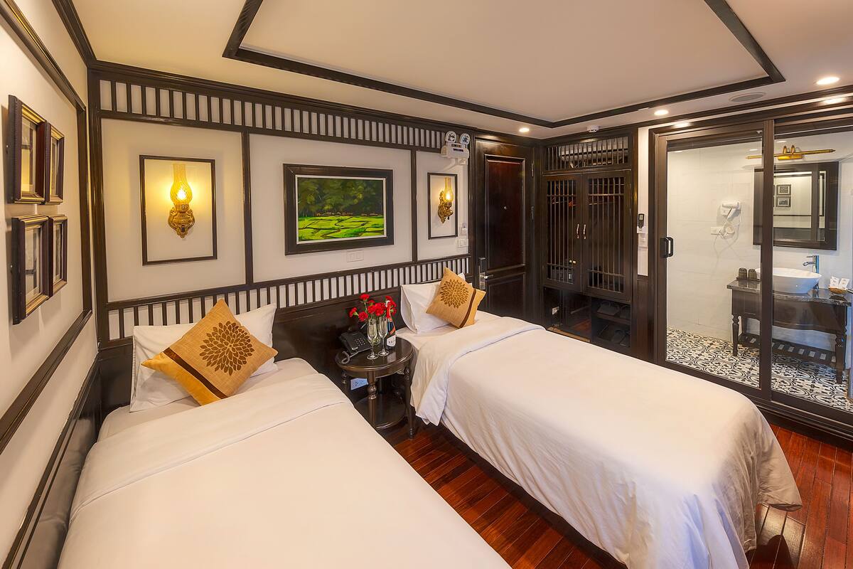 A hotel room with two beds, a small table with a phone and flowers, and a bathroom visible through a glass door.