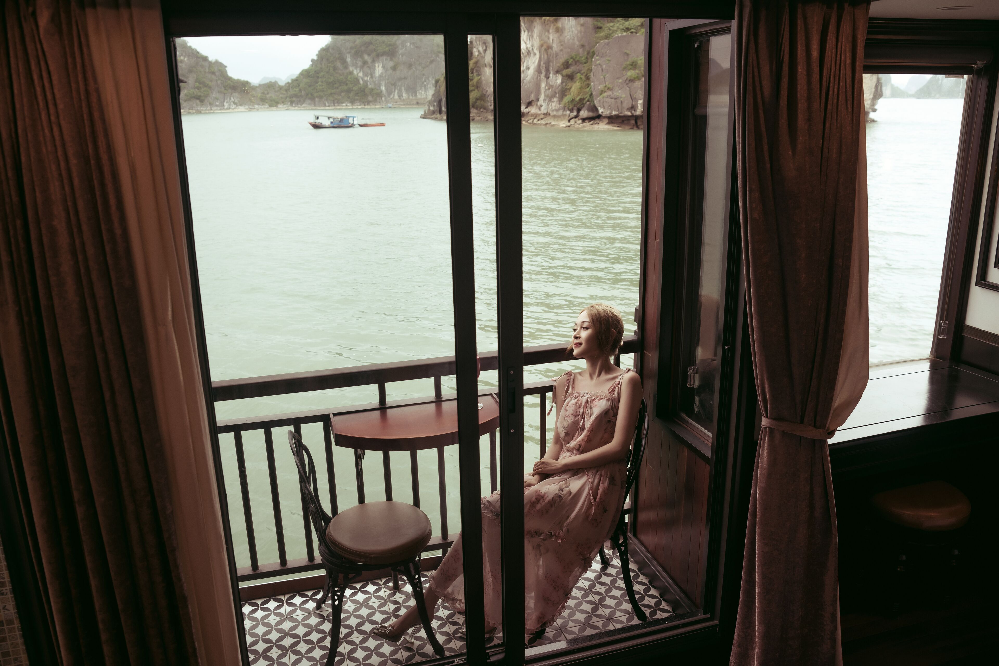 A room with a view of a calm body of water and boats, a small round table, and a chair.