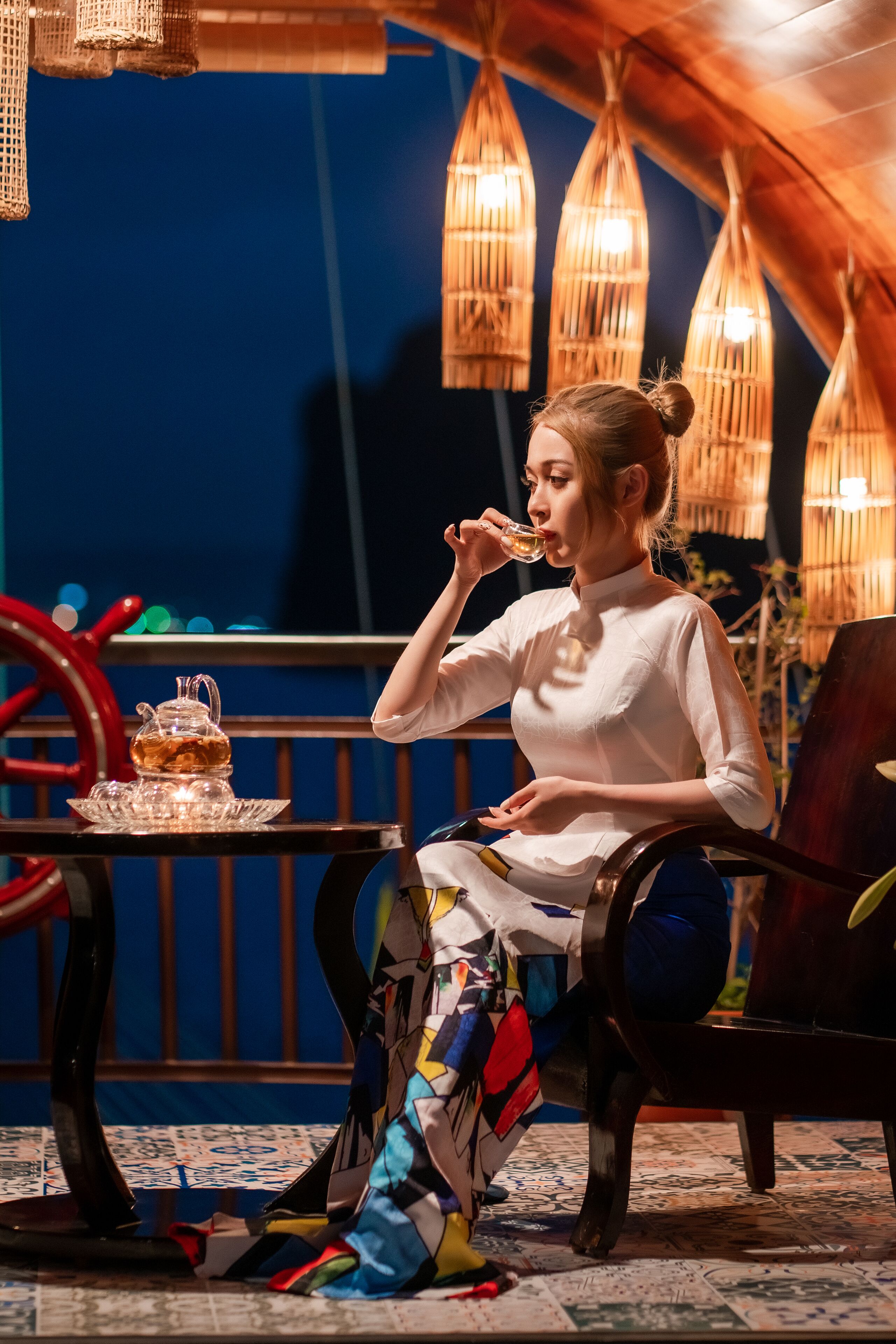 Une personne est assise à une table devant une théière et une tasse, dans une pièce dont le sol est décoré de motifs et où l’on aperçoit, au fond, un gouvernail de navire.