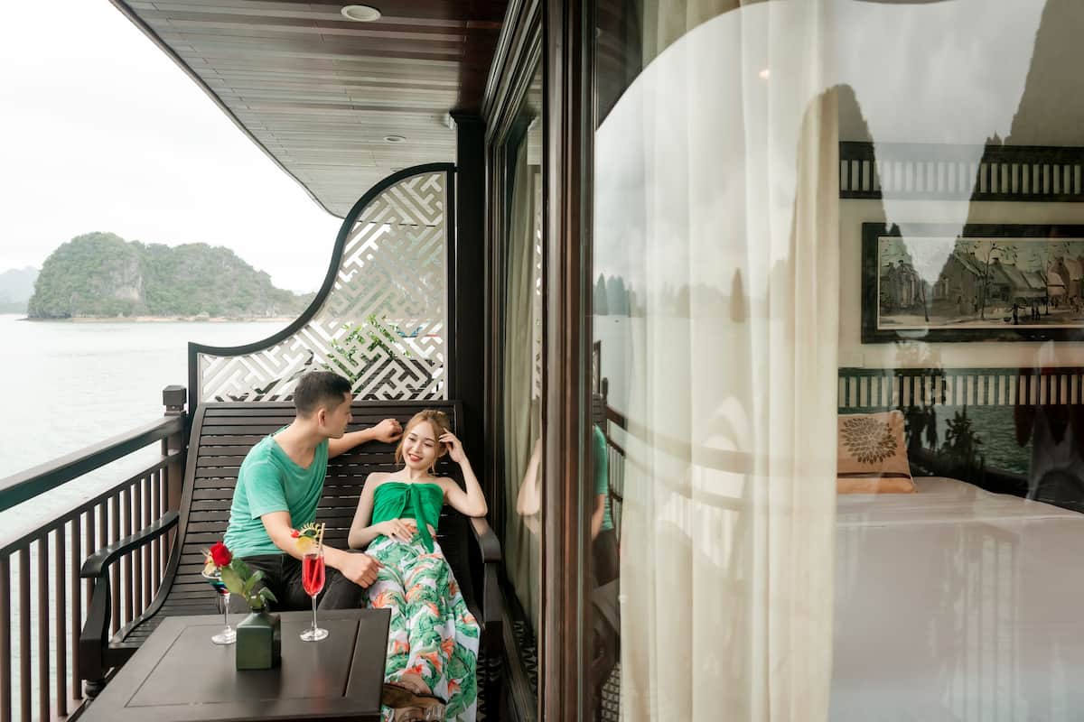 A balcony with a view of the sea, a table with drinks, and a bed with a pillow and a painting.