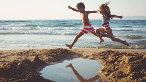 Two children jumping on a sandy beach with a puddle and ocean waves in the background.