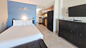 A bedroom with a bed, a dresser, and a TV. There is a kitchen in the background with wooden cabinets and a refrigerator.