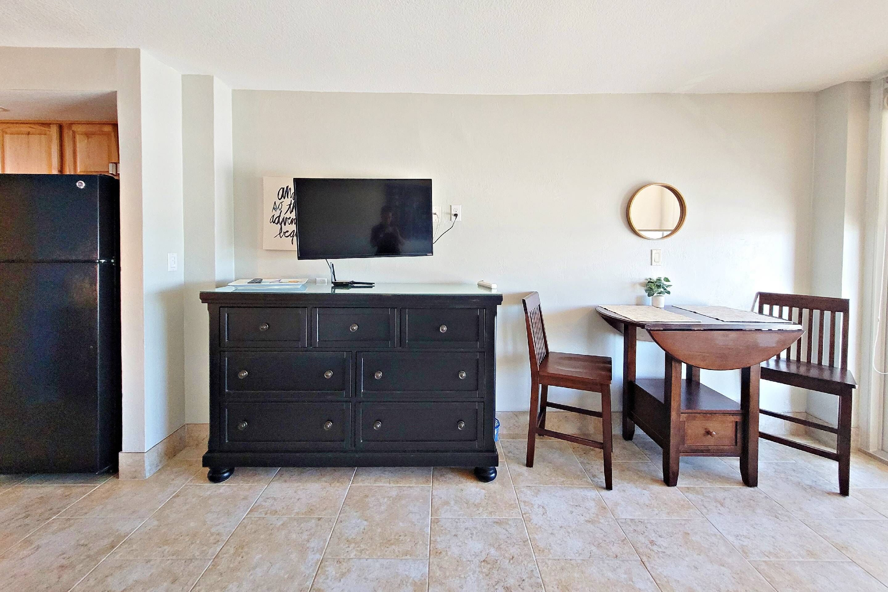 A kitchen with a black refrigerator, a wooden cabinet, a dining table with chairs, and a flat-screen TV mounted on the wall.
