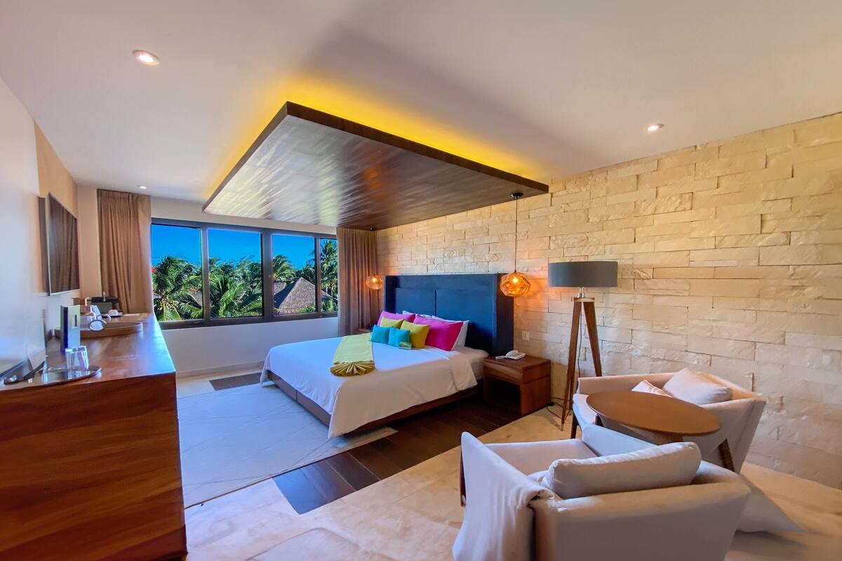 A modern hotel room with a large bed, a desk, a sofa, and a view of palm trees.