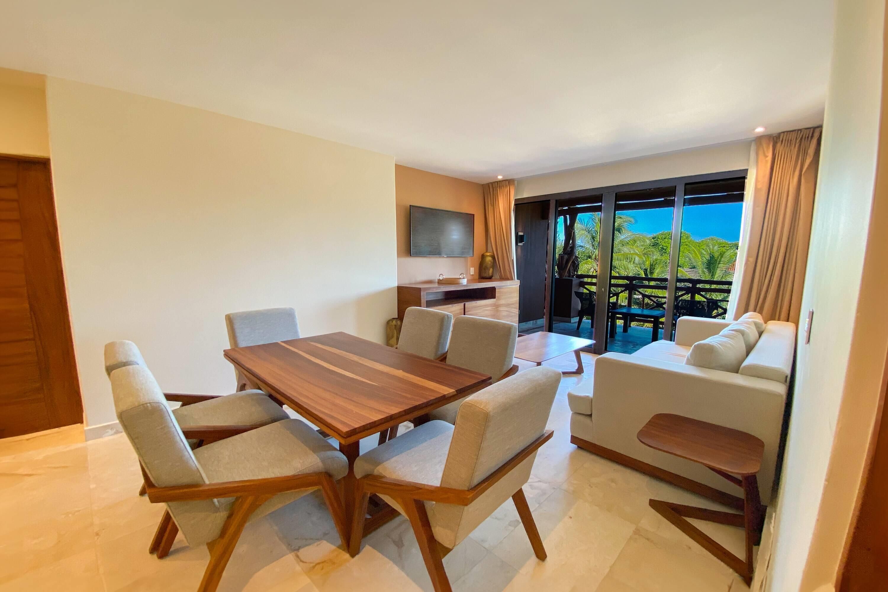 Uma sala de estar moderna com uma mesa de jantar de madeira, sofás em tons claros e uma TV fixada na parede.