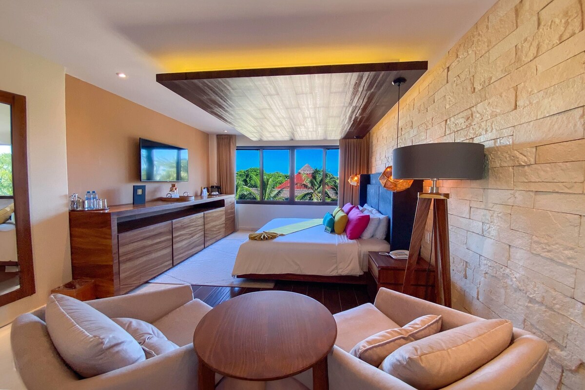 A modern hotel room with a bed, seating area, TV, and a view of the outdoors.