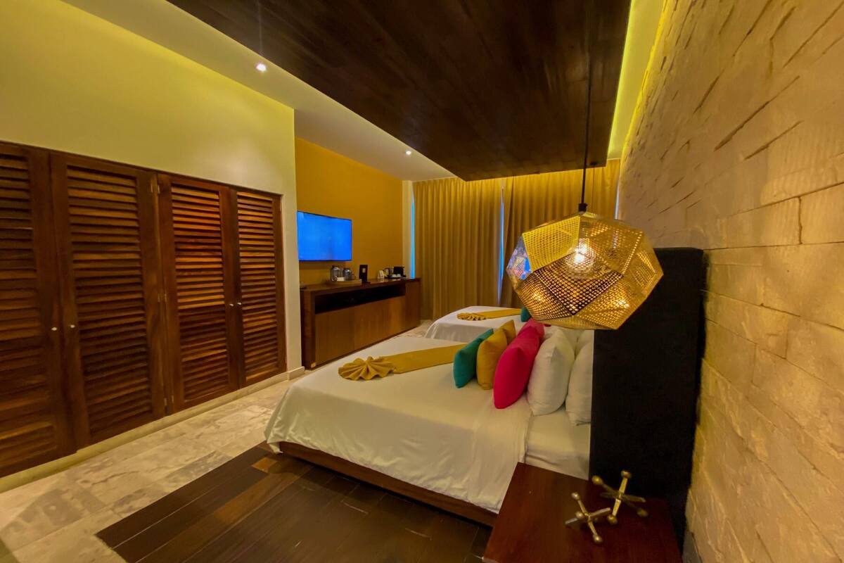 A hotel room with a bed, wooden wardrobe, a desk with a flat-screen TV, and a decorative hanging light fixture.
