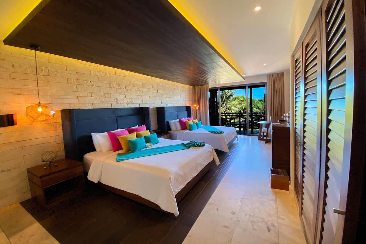 A hotel room with two beds, a brick wall, a wooden ceiling, and a view of palm trees.