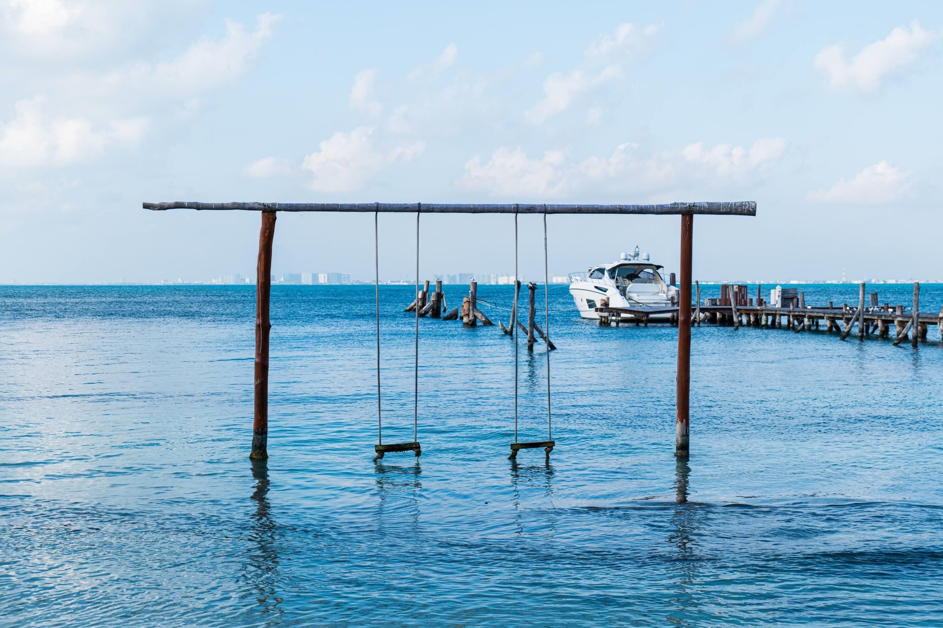 A pier with a swing set over water, a yacht docked, and a city skyline in the background.