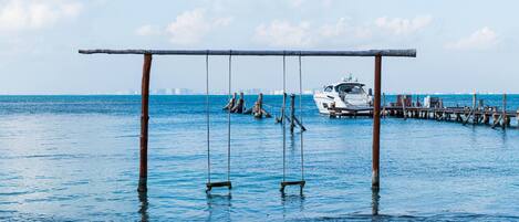 A pier with a swing set over water, a yacht docked, and a city skyline in the background.