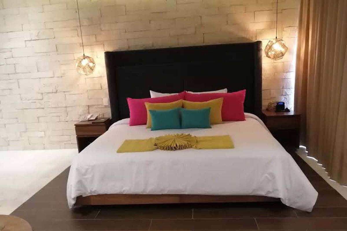 A large bed with a white bedspread, colorful pillows, and a decorative yellow towel. There are two bedside tables with lamps, and a brick-patterned wall behind the bed.