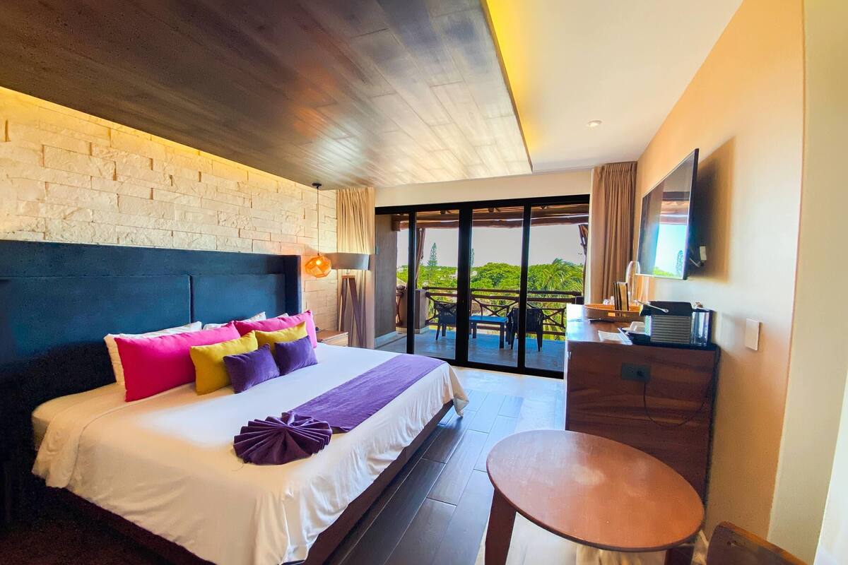 A hotel room with a large bed, a desk, a television, and a balcony with a view of greenery.