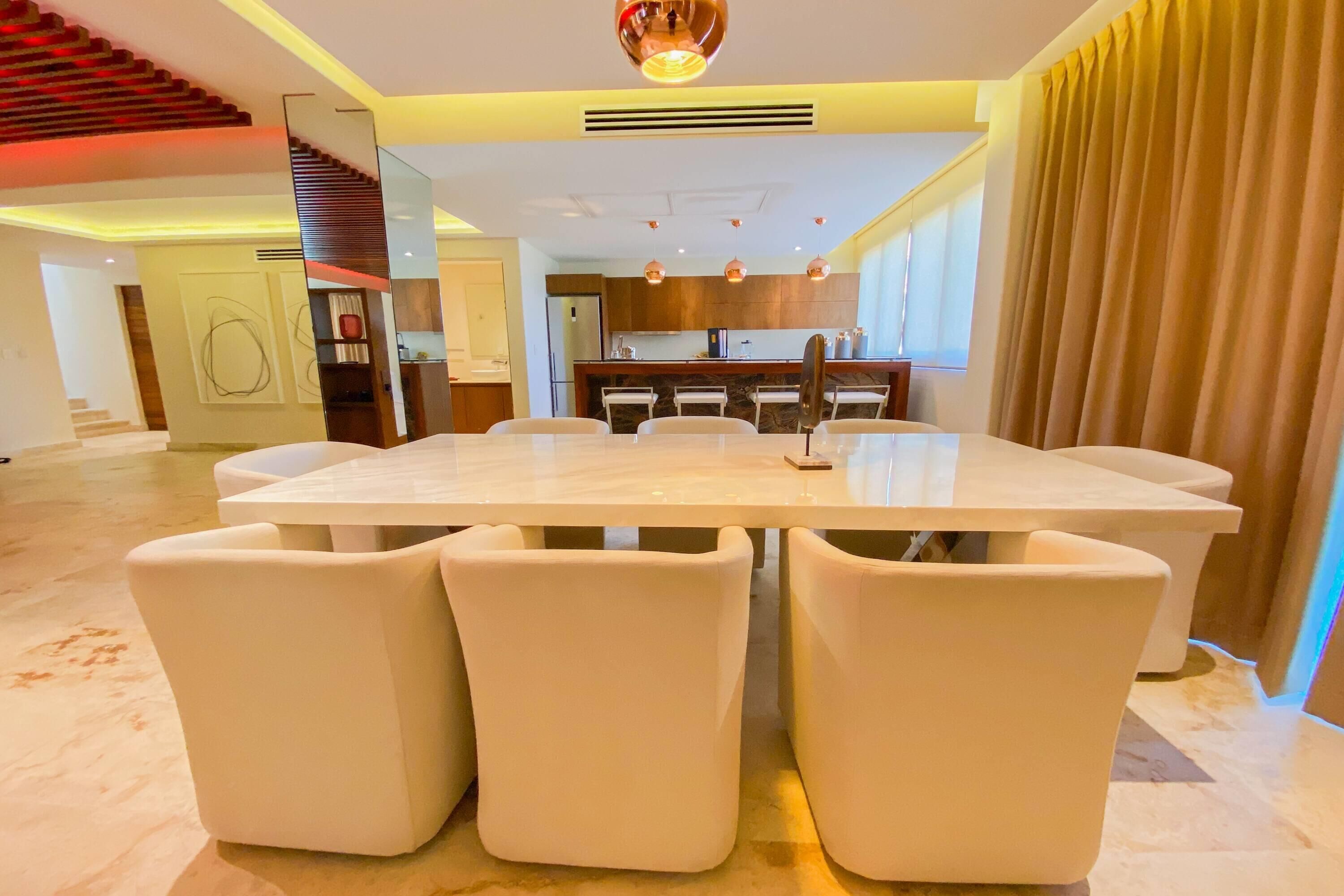 Un comedor moderno con una mesa de mármol y sillas blancas, una cocina al fondo y una escalera que sube a un nivel superior.