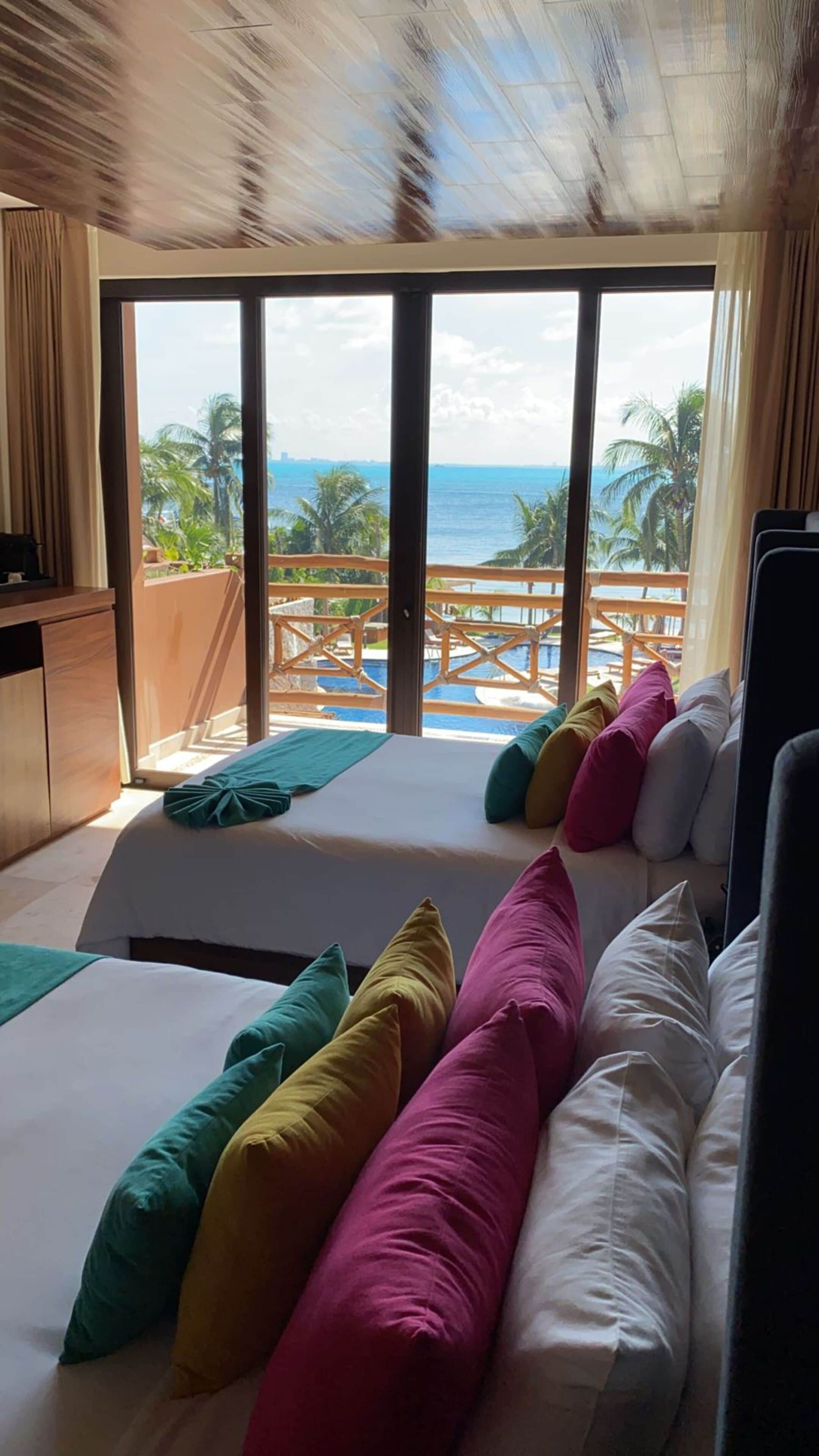 Une chambre d’hôtel avec deux lits, des oreillers colorés, une vue sur l’océan et un espace piscine visible à travers les portes vitrées.