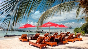 A beach with wooden lounge chairs and red umbrellas, a pier extending into the water, and palm trees. - Esplendor by Wyndham Isla Mujeres Boutique All Inclusive (Isla Mujeres)