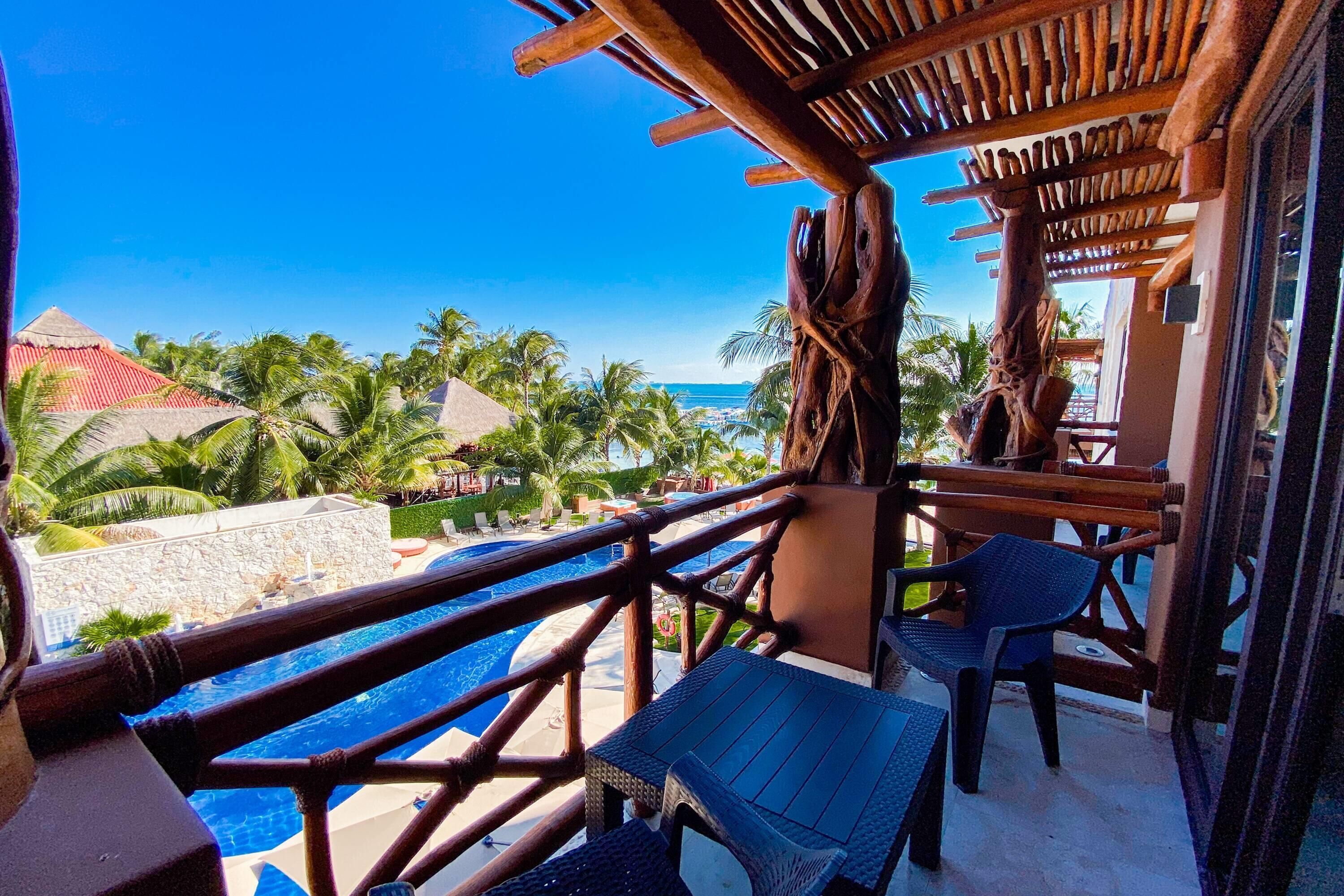 A balcony with a view of a pool and palm trees, wooden furniture, and a wooden pergola.