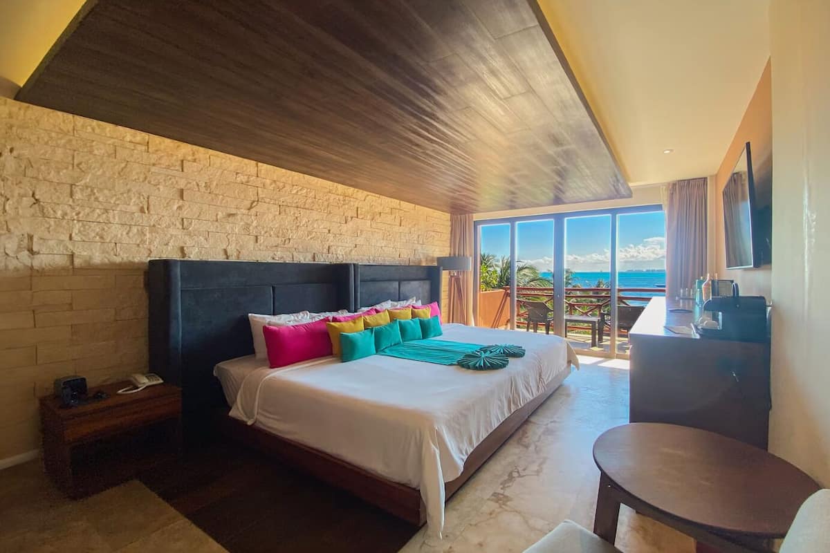 A bedroom with a large bed, a wooden headboard, a nightstand, a desk, and a view of the ocean.