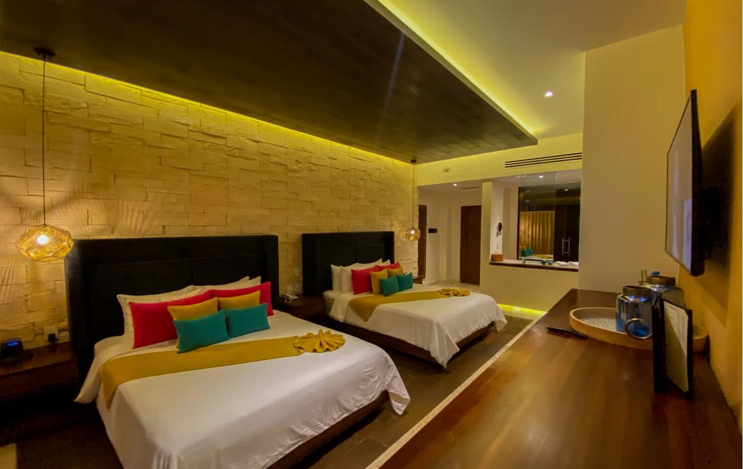 Habitación de hotel con dos camas, una pared de ladrillo, suelo de madera, un televisor y una pequeña mesa con lámpara.