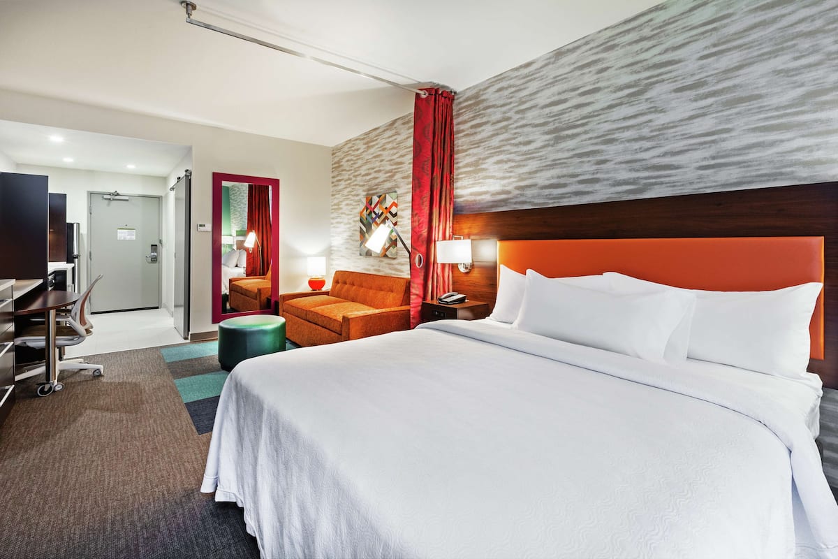 A hotel room with a large bed, a desk with a chair, a small table with a lamp, and a red curtain.