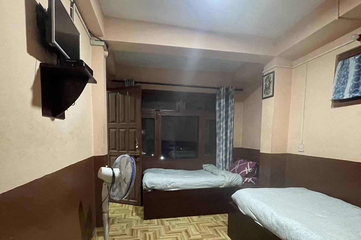Habitación de hotel con dos camas individuales, un ventilador de pie, un televisor montado en la pared y una puerta que da a un balcón.