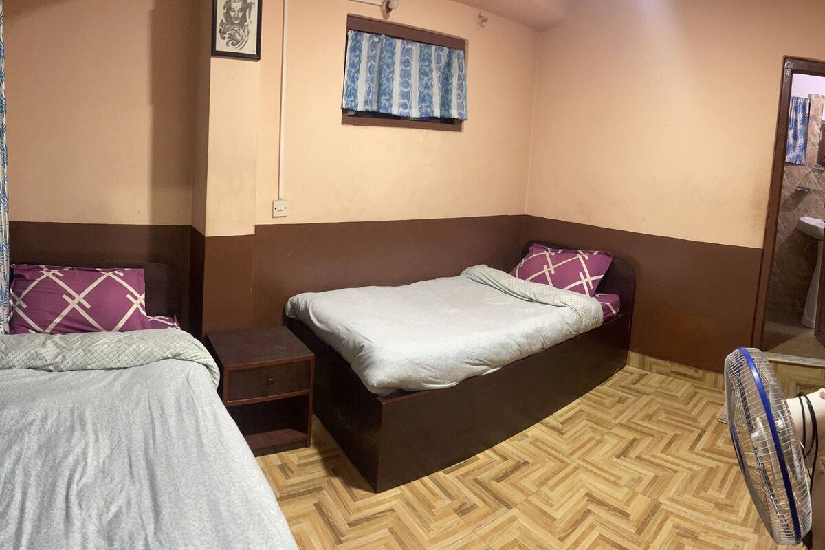 Habitación pequeña y funcional con dos camas individuales, una mesita de noche, un ventilador de pared y una ventana con cortinas decoradas.