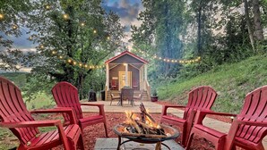 A cozy outdoor seating area with red chairs and a fire pit, overlooking a wooden cabin with string lights. - Little Red Roof, a tiny house in the GA mountains! (Clayton)