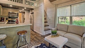 A kitchen with a microwave, stove, and sink, a bar area with stools, a staircase, and a sofa with a small table and plant. - Little Red Roof, a tiny house in the GA mountains! (Clayton)