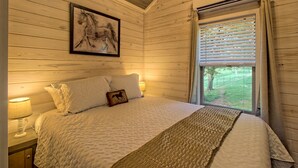 A bedroom with a bed, a nightstand, a lamp, a window with blinds, and a framed picture on the wall. - Little Red Roof, a tiny house in the GA mountains! (Clayton)