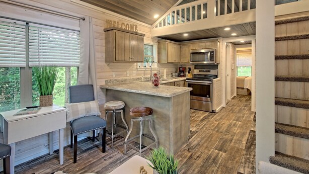 A spacious kitchen with a central island, wooden cabinets, and a "WELCOME" sign. - Little Red Roof, a tiny house in the GA mountains! (Clayton)