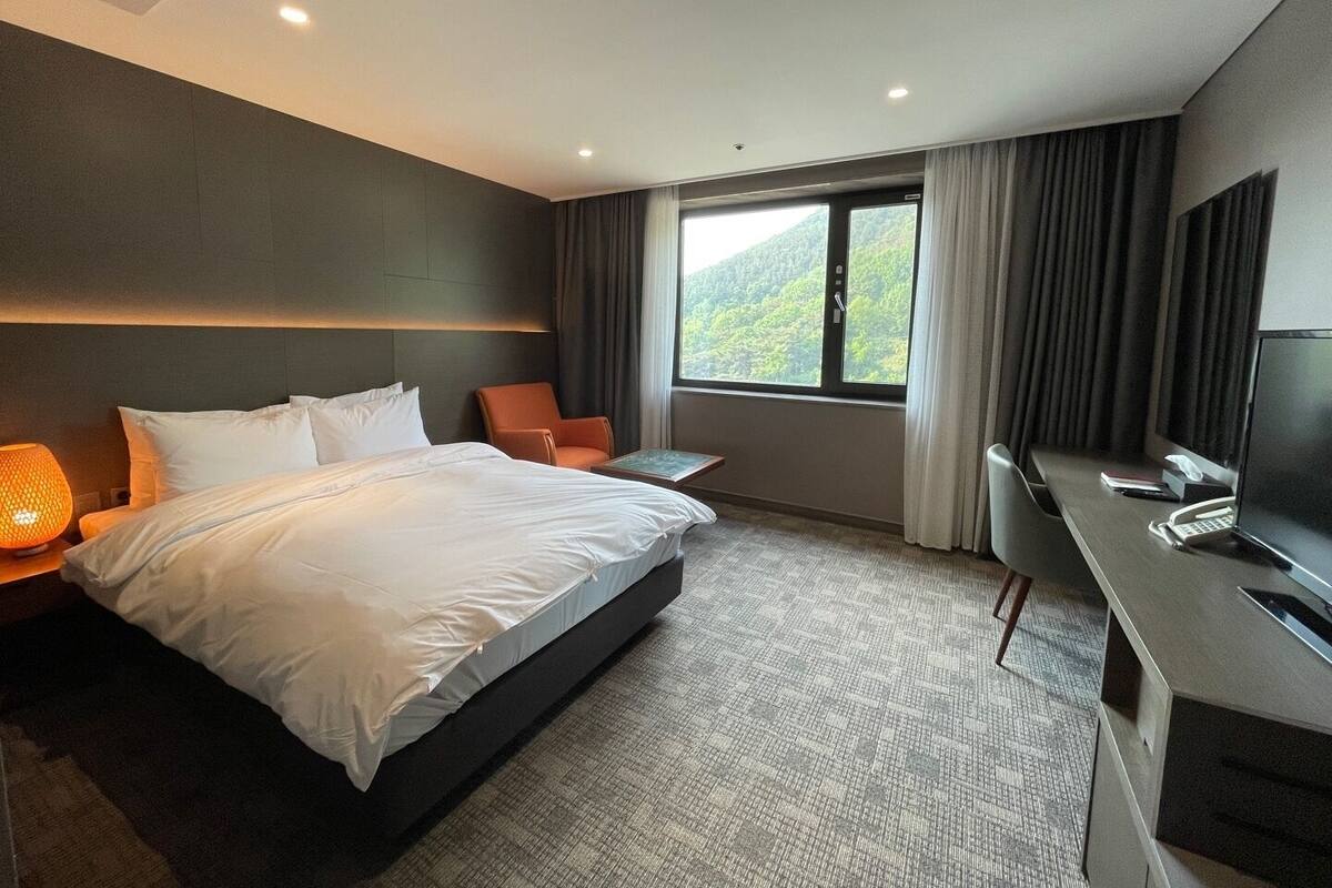 A modern hotel room with a large bed, a desk, a chair, a TV, and a window with a view of greenery.